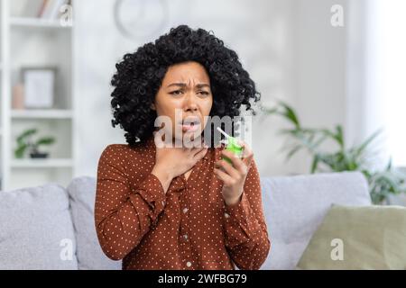 Nahaufnahme einer jungen afroamerikanischen Frau, die ihren Hals zu Hause mit einem medizinischen Spray behandelte, zu Hause auf der Couch saß und ihren Hals hielt und starke Schmerzen spürte Stockfoto