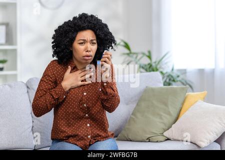 Eine junge Afroamerikanerin, die an Asthma leidet, sitzt zu Hause auf der Couch. Hält seine Brust und benutzt einen Inhalator. Stockfoto