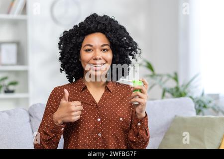 Nahaufnahme Porträt einer jungen afroamerikanischen Frau, die zu Hause auf dem Sofa sitzt, medizinisches Kehlspray hält, Superfinger zeigt, lächelt und in die Kamera blickt. Stockfoto
