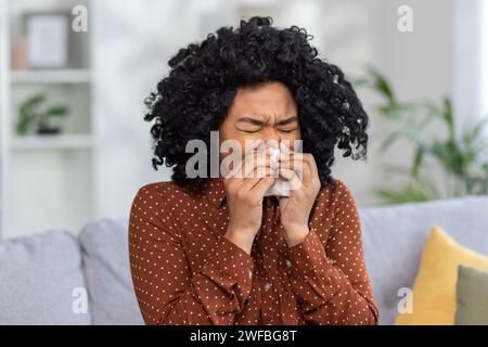 Junge Frau niest in Gewebe zu Hause, zeigt Symptome einer Erkältung oder Allergien, mit einem verschwommenen inneren Hintergrund. Stockfoto