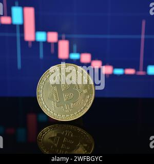 Foto von goldenem Bitcoin auf schwarzer reflektierender Oberfläche und Kerzendiagramm mit Preisverfall oder -Rückgang im Hintergrund Stockfoto