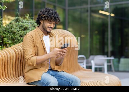 Ein lächelnder Indianer sitzt auf einer Bank in einer Stadtstraße und benutzt das Telefon. Stockfoto