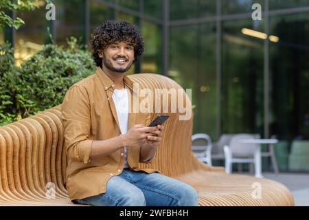 Porträt eines jungen Indianers, der auf einen Termin wartet, auf einer Bank auf der Straße sitzt, ruht, ein Telefon hält und die Kamera anlächelt. Stockfoto