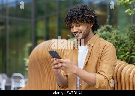 Nahaufnahme eines jungen, lächelnden Indianers, der auf einer Bank in der Nähe eines Gebäudes sitzt und das Telefon benutzt. Stockfoto