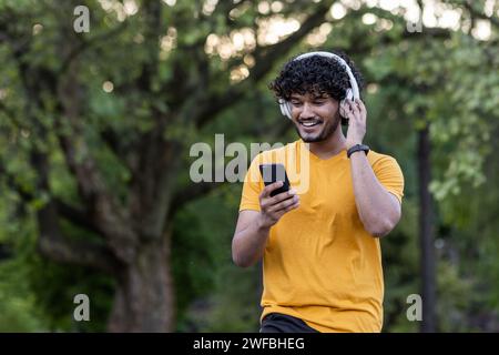 Indischer lächelnder junger Mann, der im Park Sport treibt und joggt, Kopfhörer trägt und ein Handy benutzt. Stockfoto