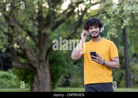 Porträt eines lächelnden jungen Indianers, der im Park mit Kopfhörern steht und das Telefon benutzt, joggt und Sport treibt. Er schaut mit einem Lächeln in die Kamera. Stockfoto