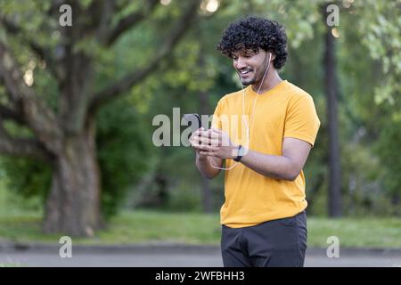 Ein junger indischer Läufer mit lockigen Haaren lächelt, während er in einem grünen Park sein Handy mit Sportbekleidung und Ohrhörern überprüft. Stockfoto