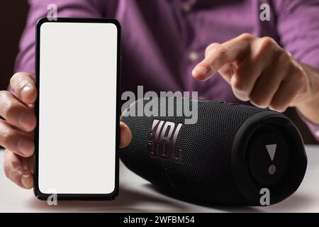 Dnioro Ukraine. 27. januar 2023: Mann hält Smartphone mit weißem Bildschirm und verwendet tragbaren Musiklautsprecher JBL Charge 5. Modell. Stockfoto