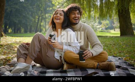 Glückliches lächelndes multiethnisches verheiratetes Paar in Liebe Mann Frau Freund Freundin beim Picknick sitzen auf Decke halten Einweg-Kaffeetassen im Park Stockfoto