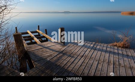 Fußgängerbrücke oder Steg bei Sonnenaufgang bei frostigem Wetter vor einem ruhigen, spiegelglatten See. Meditativer Panoramablick über das Steinhuder Meer. Stockfoto