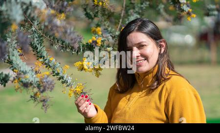 Porträt einer schönen lateinamerikanischen Frau, die fröhlich lächelt, in einer gelben Jacke gekleidet, neben einem Baum mit kleinen gelben Blüten stehend, während eines Su Stockfoto