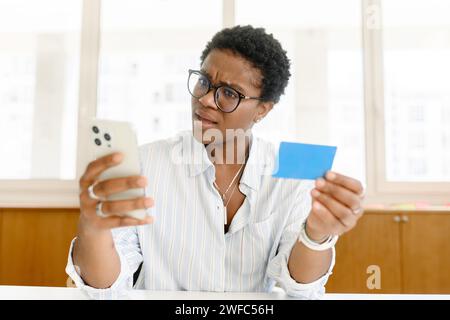 Verwirrte junge afroamerikanische Frau mit kurzen Haaren, die Smartphone und Kreditkarte hält, Schulden hat, weibliche Büroangestellte oder Freiberuflerin kein Gehalt erhalten hat, Kontostand mit traurigem Gesicht überprüft Stockfoto