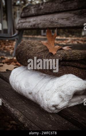 Weiße und braune Strickpullover auf einer hölzernen Stadtbank unter gefallenen Eichenblättern Stockfoto