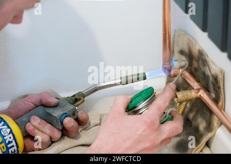 Ein Klempner, der Kupferrohre mit einem Lötbrenner erhitzt. Der Heizungsingenieur verbindet zwei Rohre mit geschmolzenem Löt über eine Winkelverbindung. Stockfoto