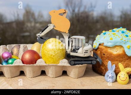 Grauer kleiner Spielzeugbagger, bunte Eier, Souvenirhasen, Osterkuchen. Osterferienkonzept für Bauunternehmen. Osterkomposition. Stockfoto