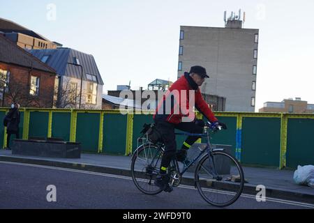 Ein weißer Radfahrer pendelt in Hackney in der frühen Wintersonne und überquert den Regent's Canal. Er trägt eine rote Jacke und eine Mütze. Stockfoto