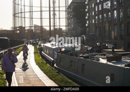 Ein Radfahrer pendelt in der frühen Morgensonne am Regent's Canal entlang. Hausboote sind auf der ganzen Seite des Kanals verankert, wo die Menschen leben. Stockfoto