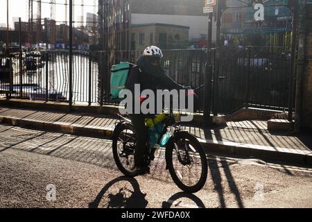 Ein Deliveroo-Mann fährt sein Fahrrad in der frühen Wintersonne entlang des Regent's Canal. Die Sonne wirft Schatten über die Straße. Stockfoto