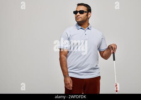 Attraktiver indischer blinder Mann in blauem T-Shirt mit Brille und Gehstock posiert vor grauem Hintergrund Stockfoto