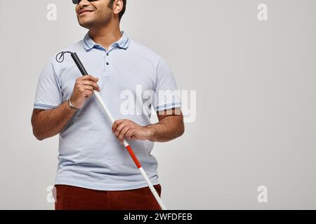 Fröhlicher indischer blinder Mann in blauem T-Shirt mit Brille und Gehstock posiert vor grauem Hintergrund Stockfoto