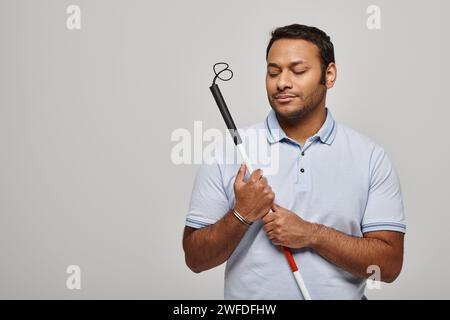 Fröhlicher indischer behinderter Mann in blauem T-Shirt, der einen Wanderstock hält und vor grauem Hintergrund posiert Stockfoto