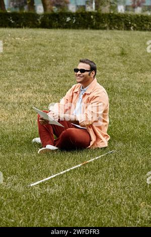 Fröhlicher indischer Blinder in orangefarbener, lebhafter Jacke, der mit Brille auf Gras sitzt und den brailleschrift liest Stockfoto