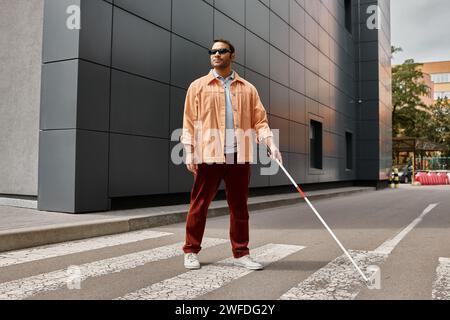 Hübscher indischer Blinder in orangefarbener Jacke mit Brille und Stock mit grauer Wand vor Hintergrund Stockfoto