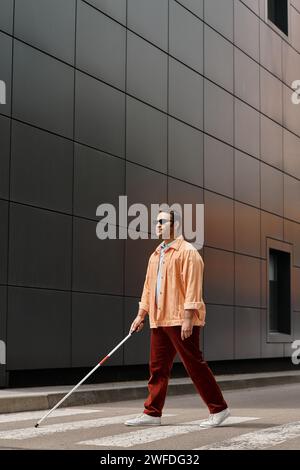 Attraktiver indischer Blindenmann in orangefarbener Jacke mit Brille und Stock mit grauer Wand im Hintergrund Stockfoto