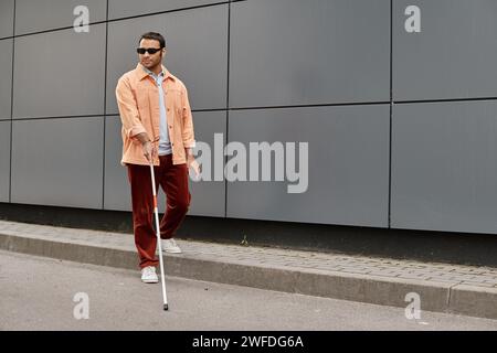 Fröhlicher indischer Blinder in orangefarbener Jacke mit Gehstock und Brille mit grauer Wand im Hintergrund Stockfoto