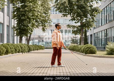 Attraktiver indischer Blindenmann in orangefarbener Jacke mit Brille und Wanderstock, der im Park spaziert Stockfoto