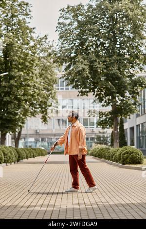 Hübscher indischer Blinder in orangefarbener Jacke mit Brille und Gehstock, der im Park spaziert Stockfoto