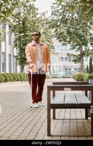 Attraktiver indischer blinder Mann mit Brille und Wanderstock in leuchtend orangefarbener Jacke, der im Park spaziert Stockfoto