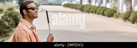 Glücklicher indischer Blinder in orangefarbener Jacke, der auf der Bank sitzt, mit Gehstock und Brille, Banner Stockfoto