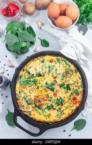 Brunch Eierfrittata mit Spinat, gebratenen roten Paprika, Pilzen, Käse und Kräutern, in gusseiserner Pfanne, vertikal, Blick von oben Stockfoto