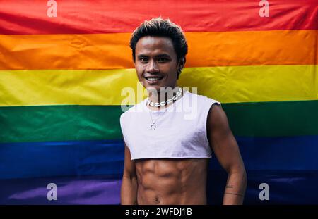 Lächelnder Mann in einem kurzen T-Shirt, der an der lgbt-Regenbogenfahne steht und in die Kamera schaut Stockfoto