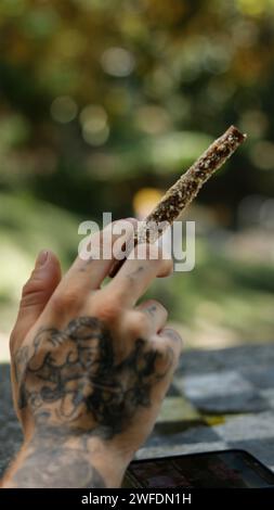 Eine tätowierte Hand, die einen Marihuana-Blunt im Outdoor-Park hält Stockfoto