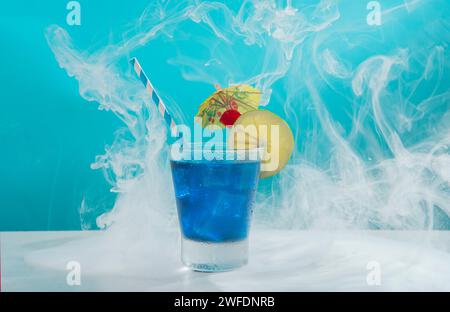Süßer Cocktail in einem kalten Glas gefüllt mit Eis garniert mit Zitronenscheiben, Kirsche, gelbem Schirm und Sorbet auf einem weißen Tisch vor hellem blau Stockfoto