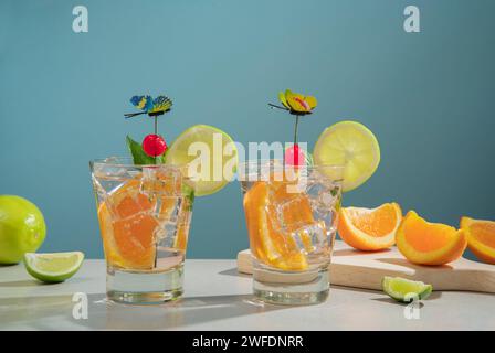 Zwei Cocktails in Glas gefüllt mit Eis und Orangenscheibe garniert mit Zitronenscheibe, Kirsche und Schmetterlingsnadel auf einem weißen Tisch mit Stücken von ora Stockfoto