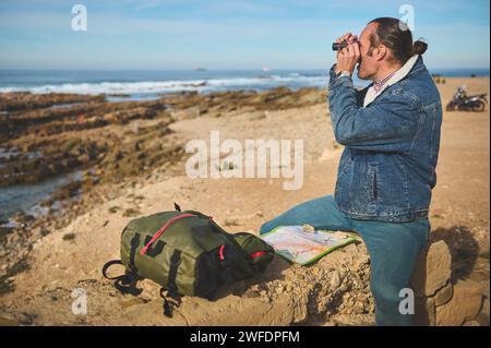 Selbstbewusster junger männlicher Abenteurer, der auf einem Felsen am Meer in der Nähe seines Rucksacks sitzt und durch sein Fernglas und Chec in die Ferne blickt Stockfoto
