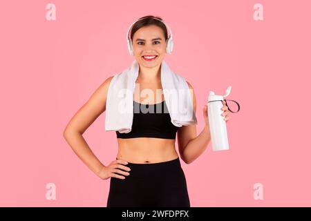 Lächelnde junge Sportfrau mit Kopfhörern und Sportbekleidung, die eine Wasserflasche hält Stockfoto