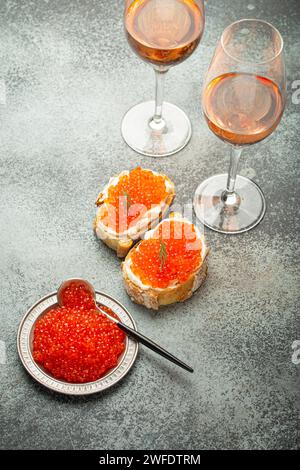 Kleine Metallplatte mit rotem Lachskaviar, zwei Kaviar Toast Canapé, zwei Gläser Champagner von oben auf grauem Betonhintergrund, festlicher Luxus Stockfoto