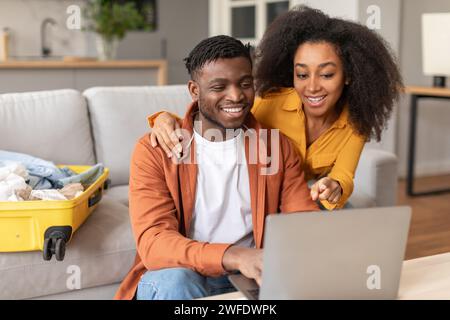 Frohe afroamerikanische Ehepartner, die ihren Laptop benutzen, um sich auf die Reise ins Haus vorzubereiten Stockfoto