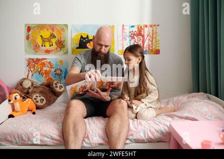 Bärtiger Mann mit offenem Comicbuch mit Bildern, die neben seiner liebenswerten kleinen Tochter auf dem Bett sitzen und ihre neuen Geschichten lesen Stockfoto