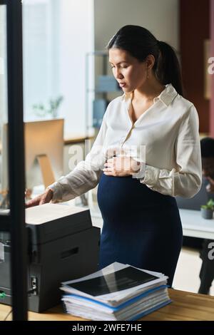Junge schwangere Büroleiterin, die vor der xerox-Maschine im Büro steht und ihren Bauch berührt, während sie Kopien von Dokumenten anfertigt Stockfoto