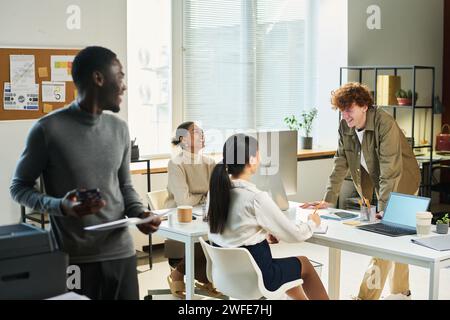 Zwei junge interkulturelle Geschäftsfrauen und ihr männlicher Kollege lachen in der Pause, während sie am Schreibtisch in einem modernen Büro sitzen Stockfoto
