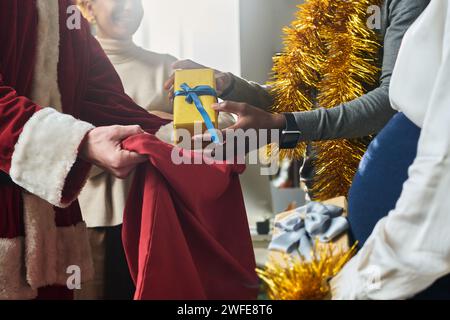 Hände eines jungen afroamerikanischen Büroangestellten, der eine Kiste über einem roten Sack hält, der von einem männlichen Kollegen gehalten wird, der Weihnachtsmannskostüm trägt Stockfoto