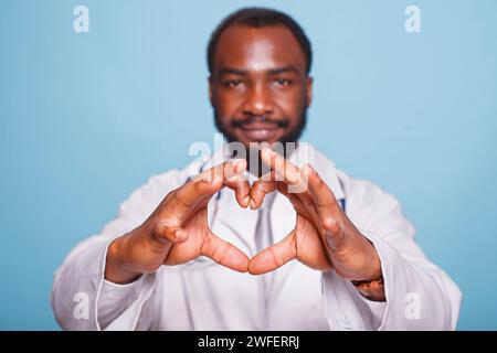 Nahaufnahme einer Herzform von Fingern eines afroamerikanischen Arztes. Schwarzer männlicher professioneller Arzt im Labormantel, der Liebesgesten zur Kamera macht. Stockfoto