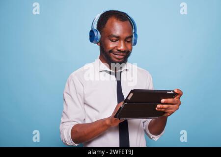 Männlicher Unternehmer, der sich mit kabellosen Kopfhörern entspannt und die Online-Show auf dem Tisch vor blauem Hintergrund betrachtet. afroamerikanischer Student, der soziale Medien auf einem digitalen Touchscreen-Gerät durchsucht. Stockfoto
