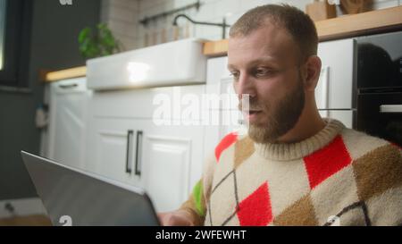 Mann, der auf dem Boden in der Küche sitzt und mit sich selbst spricht, auf einem Laptop schreibt und im Internet surft, soziale Medien nutzt und mit Freunden chatten kann Stockfoto