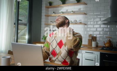 Müder Mann, der in der Küche sitzt, hat starke Schmerzen im Nacken, reibt ihn, um Muskelverspannungen zu lösen, massiert seinen Nacken und macht eine Pause von der Arbeit Stockfoto